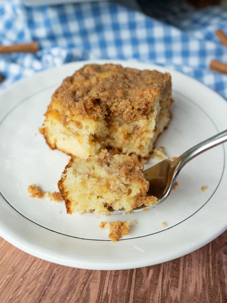 one piece of cinnamon streusel cake on a white plate that is sitting on a blue and white cloth there is a fork on the plate with a bite of cake on it on a wood countertop