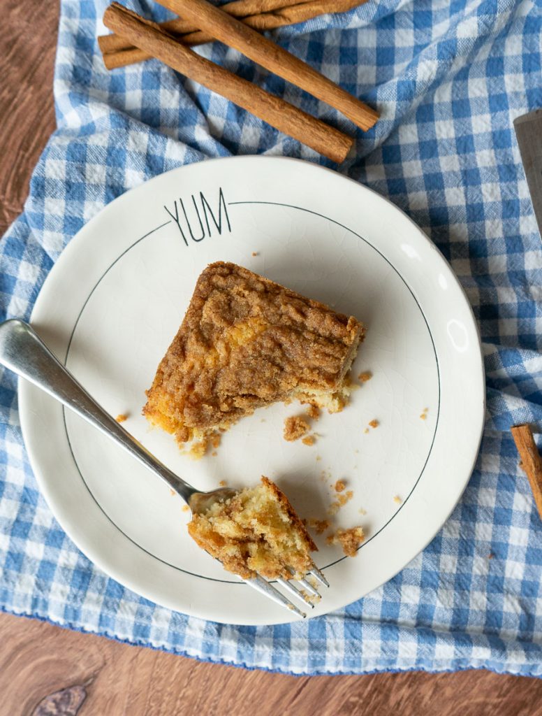 a piece of coffee cake on a white small plate half eaten with a fork next to the cake with a bite of cake on the fork plate is setting on a blue and white checked cloth there are cinnamon sticks on the cloth