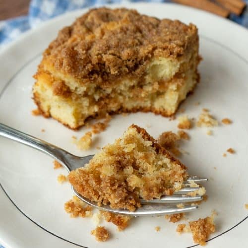 one piece of cinnamon streusel cake on a white plate that is sitting on a blue and white cloth there is a fork on the plate with a bite of cake on it on a wood countertop