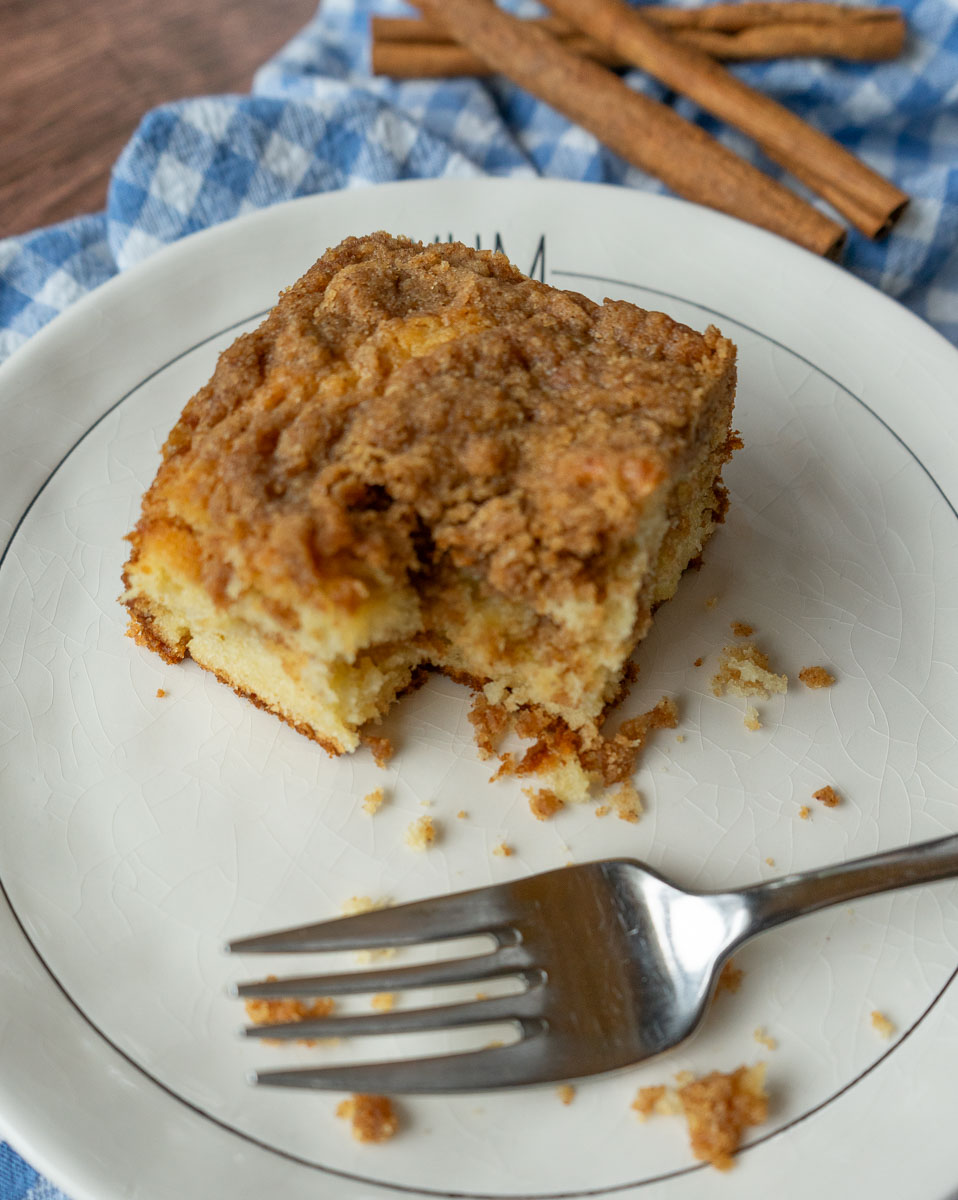 a slice of coffee cake on a white plate with one bite taken a fork is laying on the plate. plate is sitting on a blue and white checked cloth with cinnamon sticks in the background