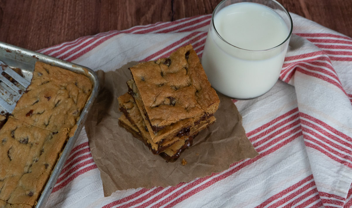 Stack of chocolate chip cookie bars on a table with a glass of milk and the pan of cookie bars off the the left side