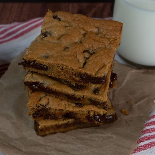 stack of chocolate chip cookie bars on a piece of brown paper with a glass of white milk next to it