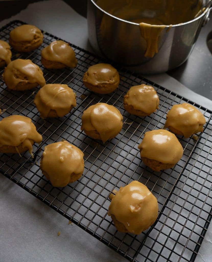 pumpkin spice cookies cooling on a wire rack with pan of frosting in the back ground