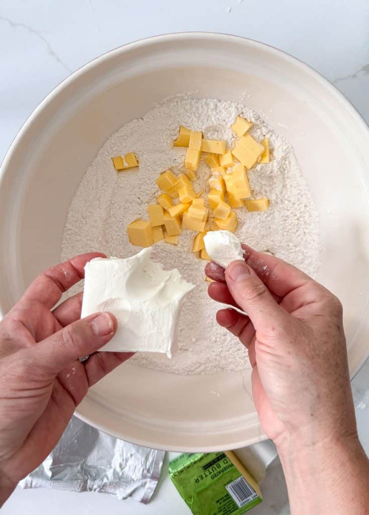 adding cold butter and cream cheese to the dry ingredients in a white bowl on a white countertop