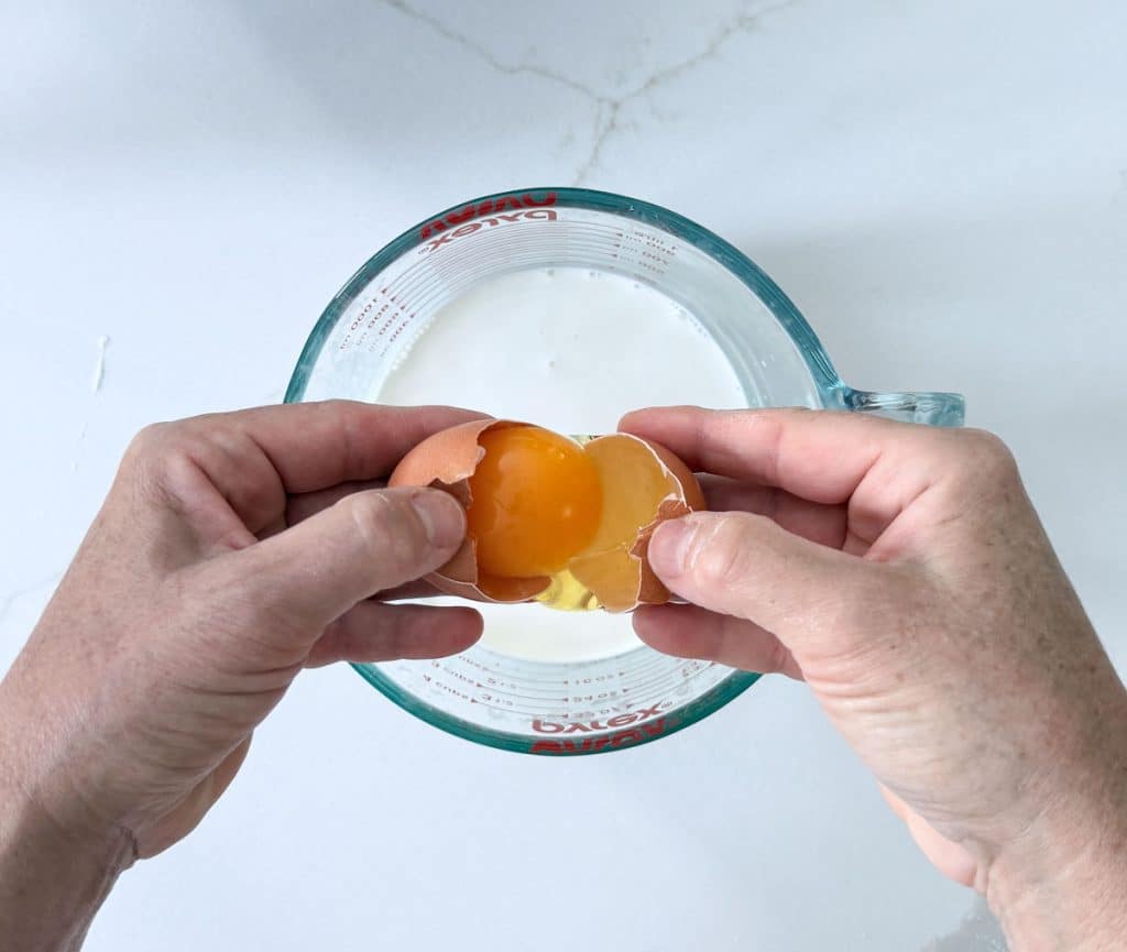 hands breaking egg into half and half that is in a glass measuring cup on a white countertop