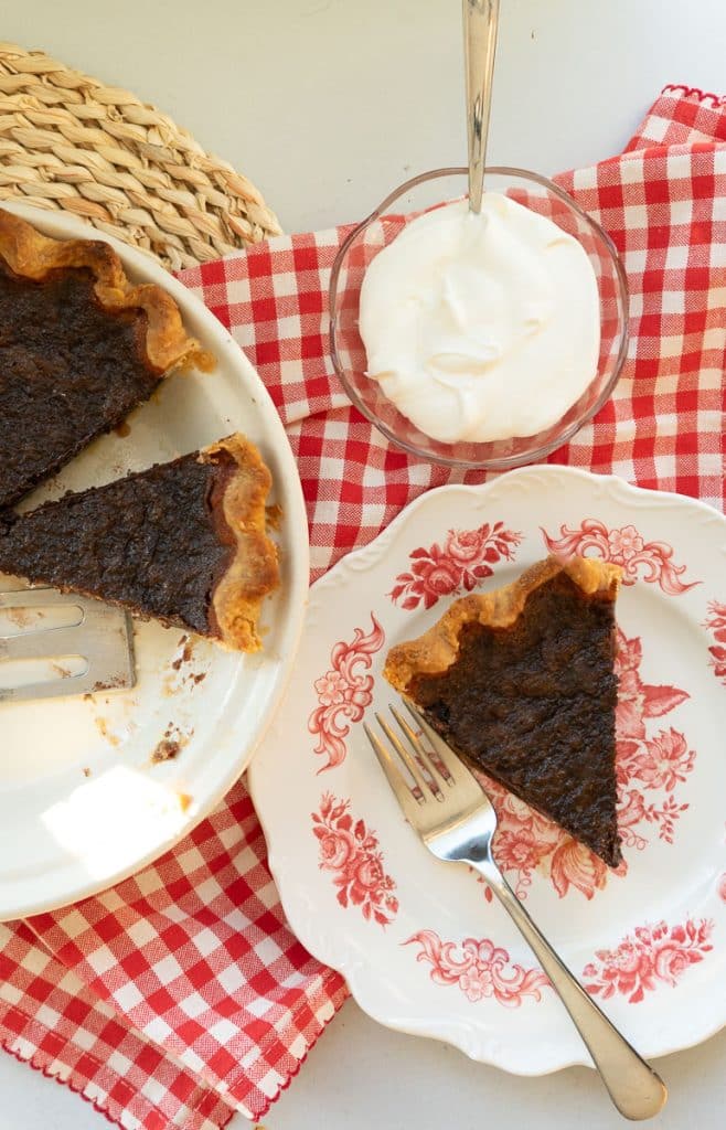 IMG_3864 chocolate pie with pieces cut out and a slice of pie on a plate with a small bowl of whipping cream on the table there is a red and white gingham napkin under the plate