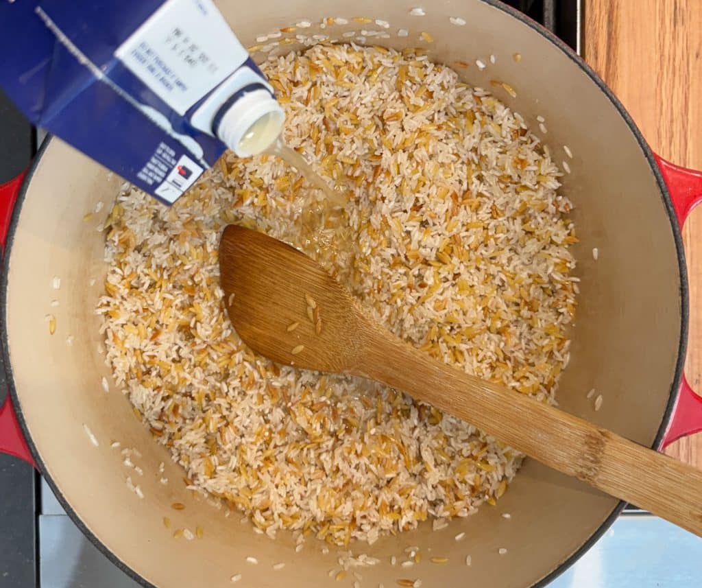 adding chicken stock to the pot with orzo pasta and rice