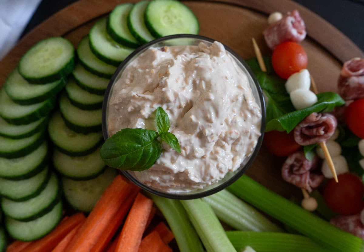 bowl of cheesy vegetable dip garnished with basil leafs. bowl is on a wood platter surrounded by fresh cut vegetables