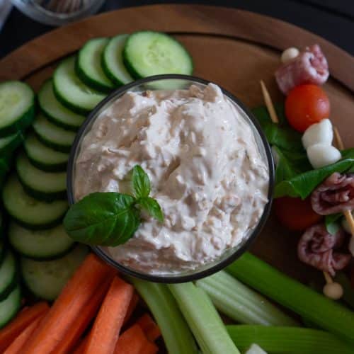 bowl of cheesy vegetable dip garnished with basil leafs. bowl is on a wood platter surrounded by fresh cut vegetables