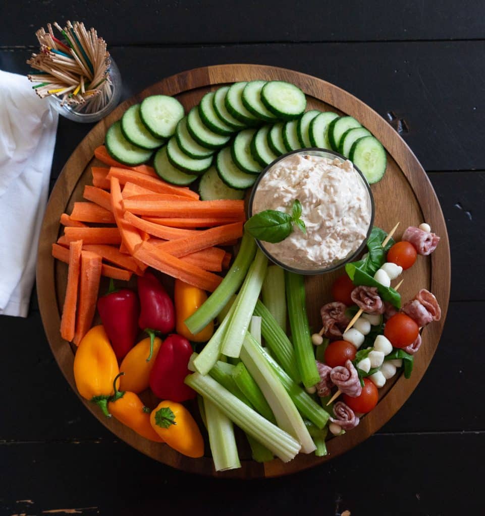 wood tray with cut vegetables and skewers of meat and cheese with a bowl of easy cheesy vegetable dip in the center