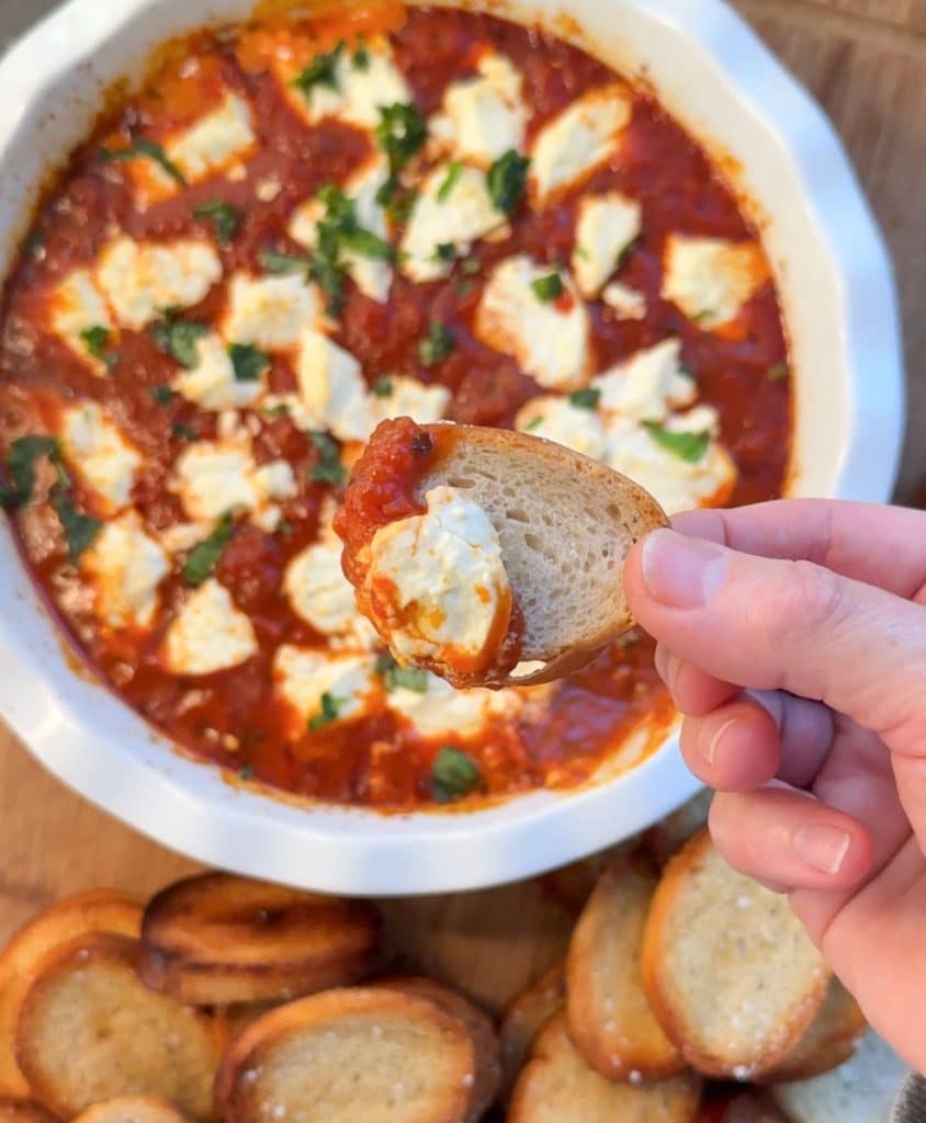 fingers holding a toast with warm Italian goat cheese dip on it over the dish of warm dip