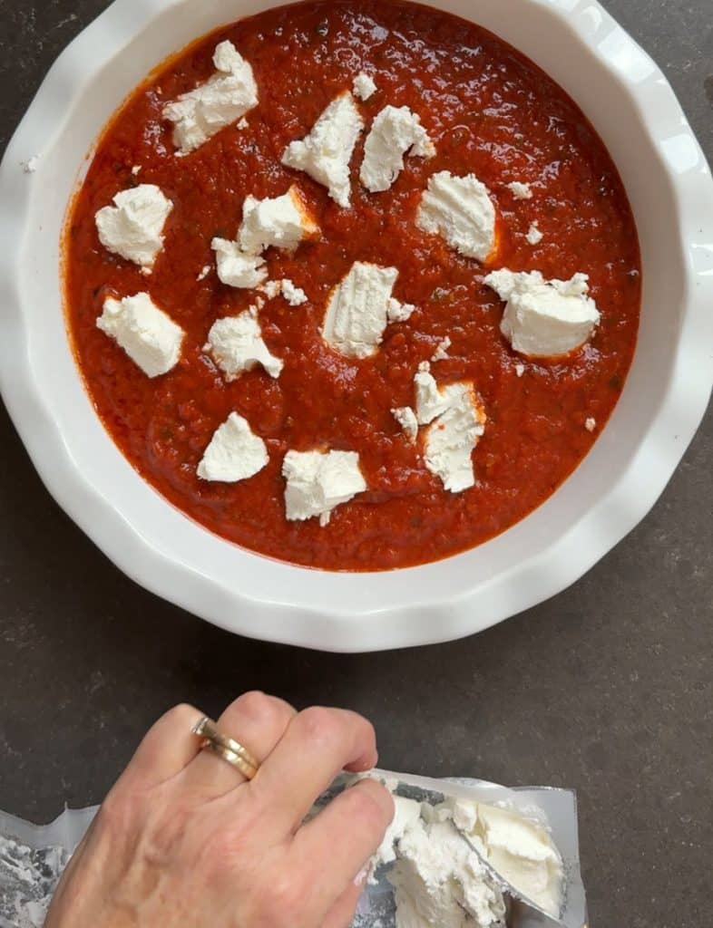 crumbling goat cheese over marinara sauce in baking dish log of goat cheese is below the baking dish on the counter