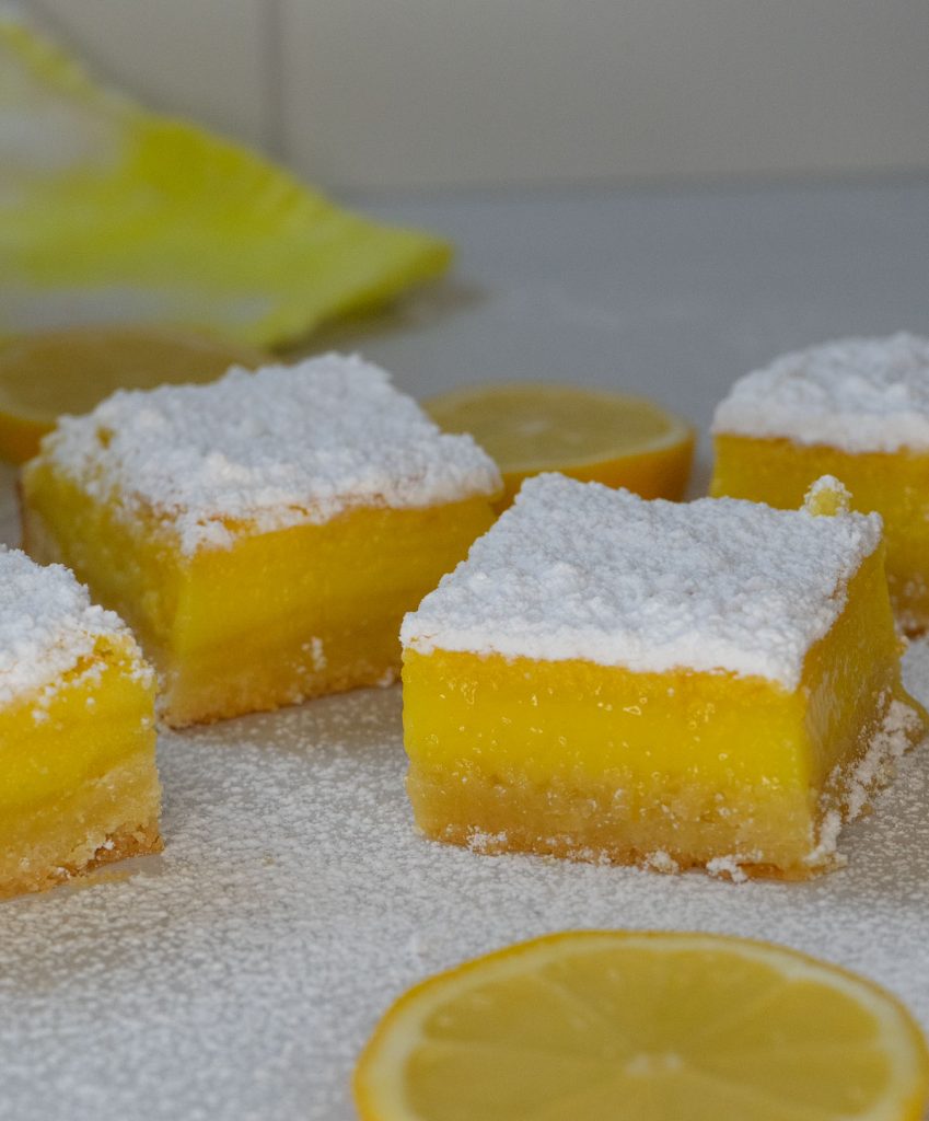 lemon bars on a grey counter with sliced lemons in the photo