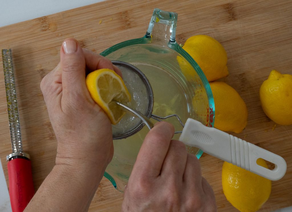 hands juicing lemon over a glass measuring cup on a wood cutting board with lemons on the board