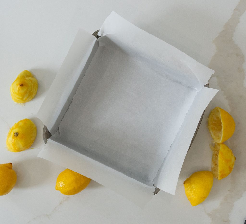 line 8x8 baking pan lined with parchment paper on a white mable countertop lemons on counter beside pan