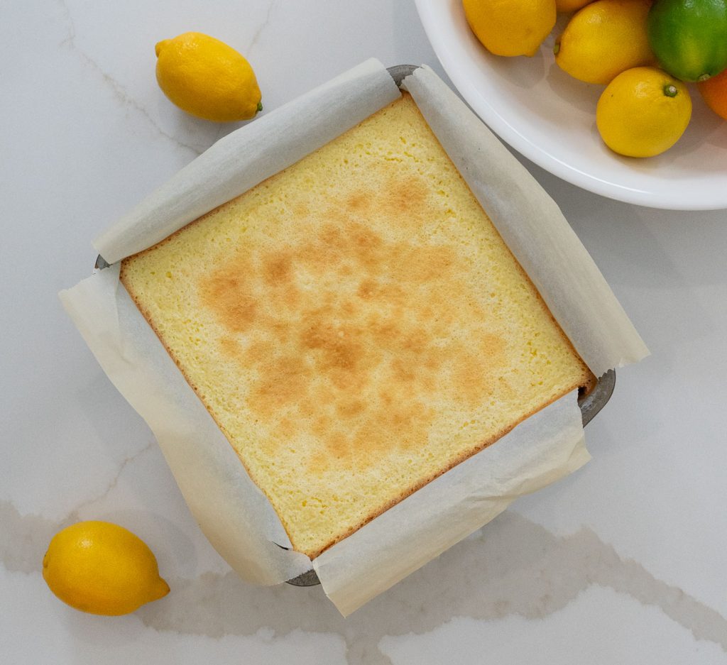 lemon bars after being baked sitting on the counter in pan with a bowl of citrus in the upper righthand corner with lemons laying around the pan
