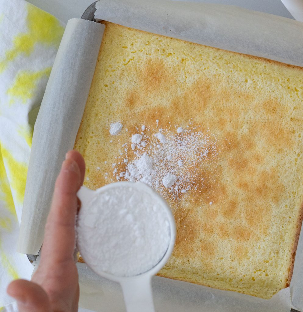 dusting lemon bars with powdered sugar. sieve full of sugar with a hand on the left tapping sugar over bars