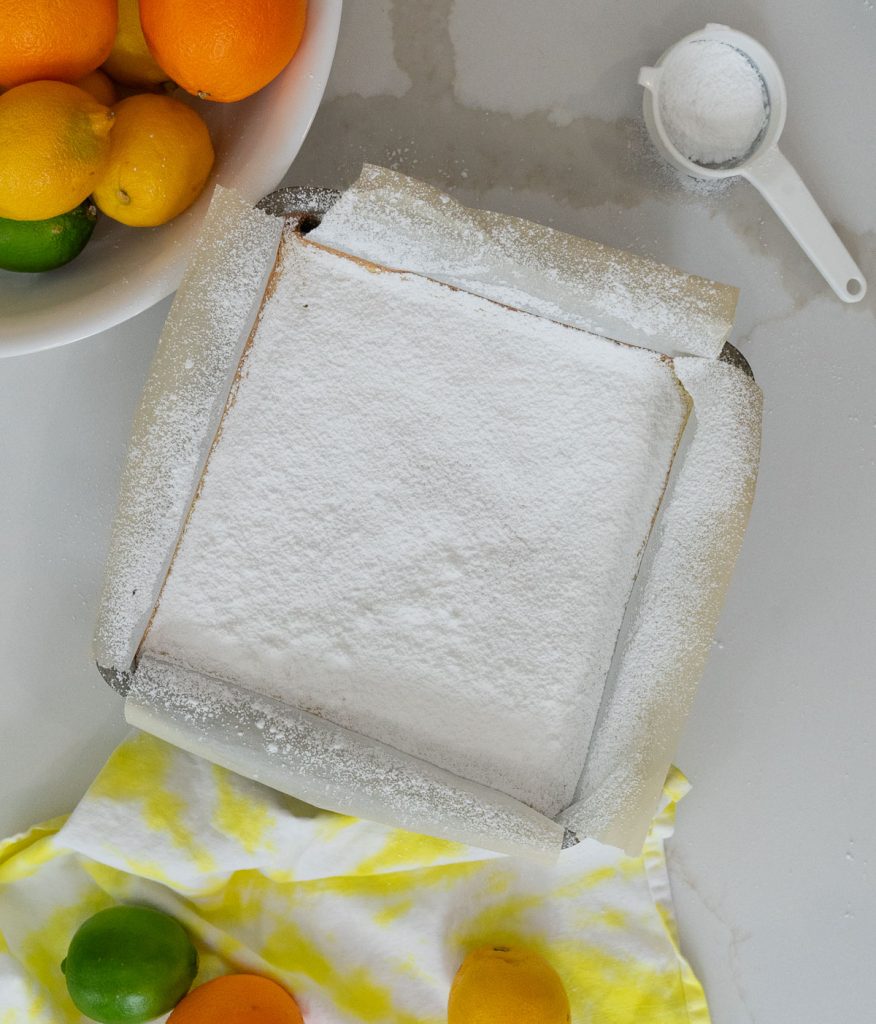lemon bars in pan with citrus around it, bars are covered in a thick dusting of powdered sugar