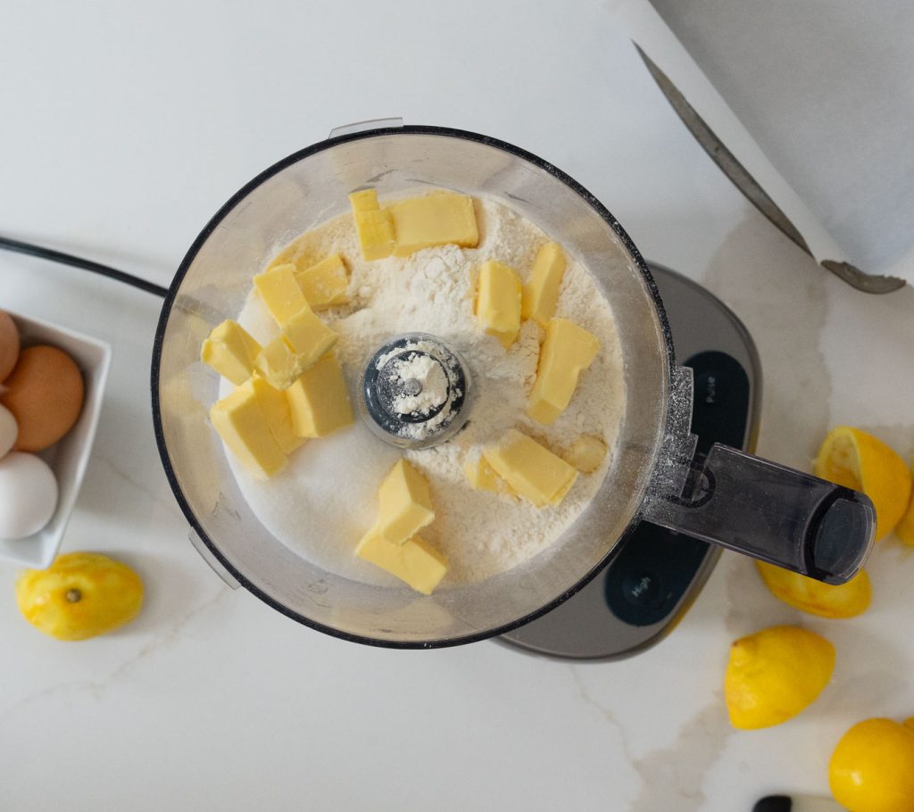 flour sugar, butter, and salt in food processor on white countertop with halves of lemons on the counter