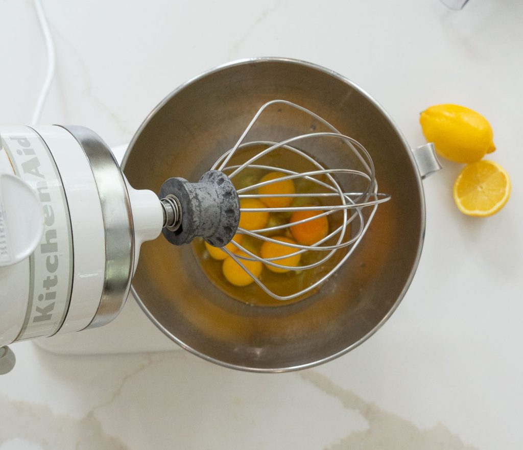 eggs in the bowl of stand mixer on a white counter with slice lemon on the counter next to the mixer