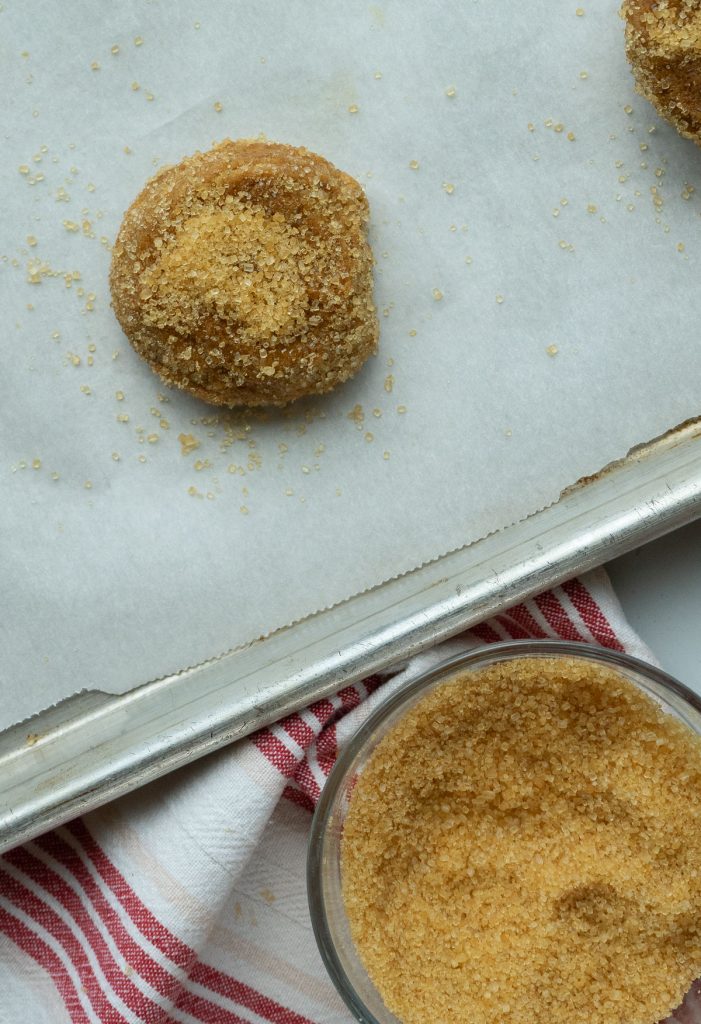 molasses cookie dough ball on a baking sheet next to bowl of turbinado sugar