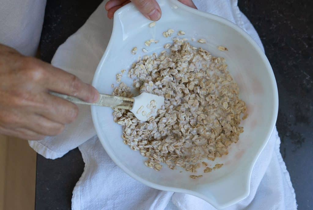 IMG_4360 bowl with oatmeal and buttermilk with a hand stirring with a small spatula in a white bowl on a black countertop . bowl is setting on a white towel