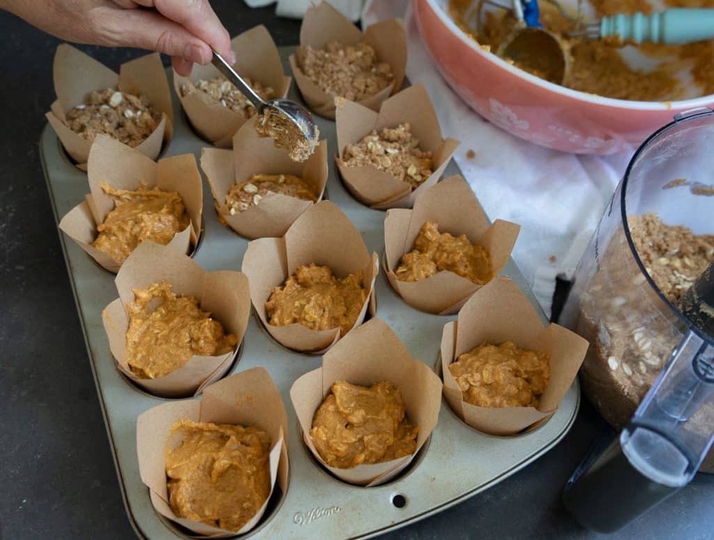 putting crumble topping on top of muffin batter in muffin cups on counter top the muffin tin is surrounded by the bowl of muffin batter and various baking utensils