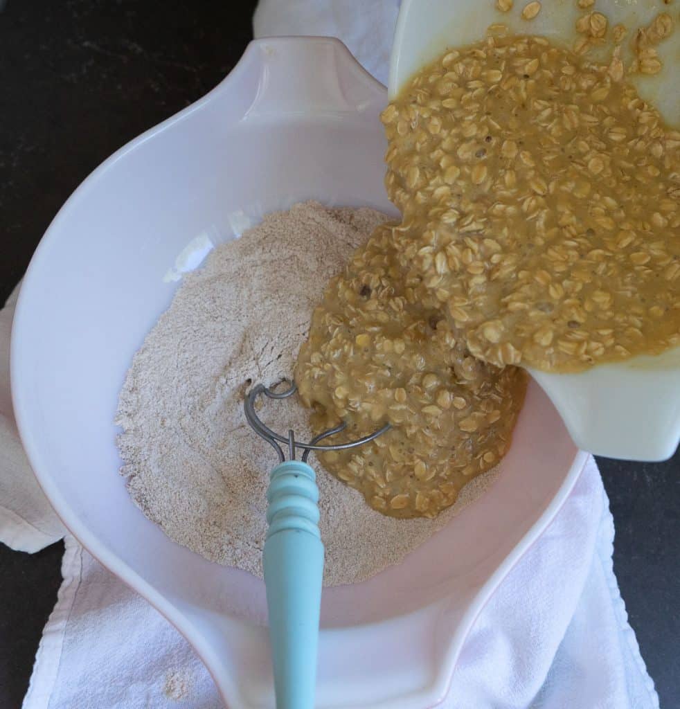 adding wet oatmeal ingredients to dry whole wheat flour ingredients. photo shows pouring into bowl, there is a bread whisk in the large bowl