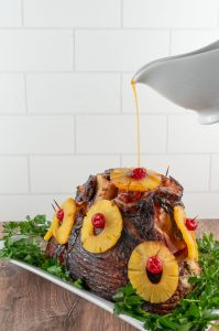ham covered with pineapple slices with maraschino cherries in the middle on a white platter surrounded by parsley and a gravy boat pouring glaze over baked ham from above