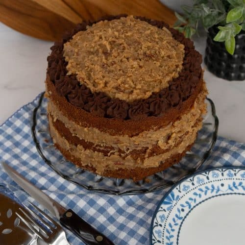 whole German Chocolate Cake with on a glass cake plate with a blue and white gingham cloth underneath cake plate, a blue trimmed plate is lower righthand corner
