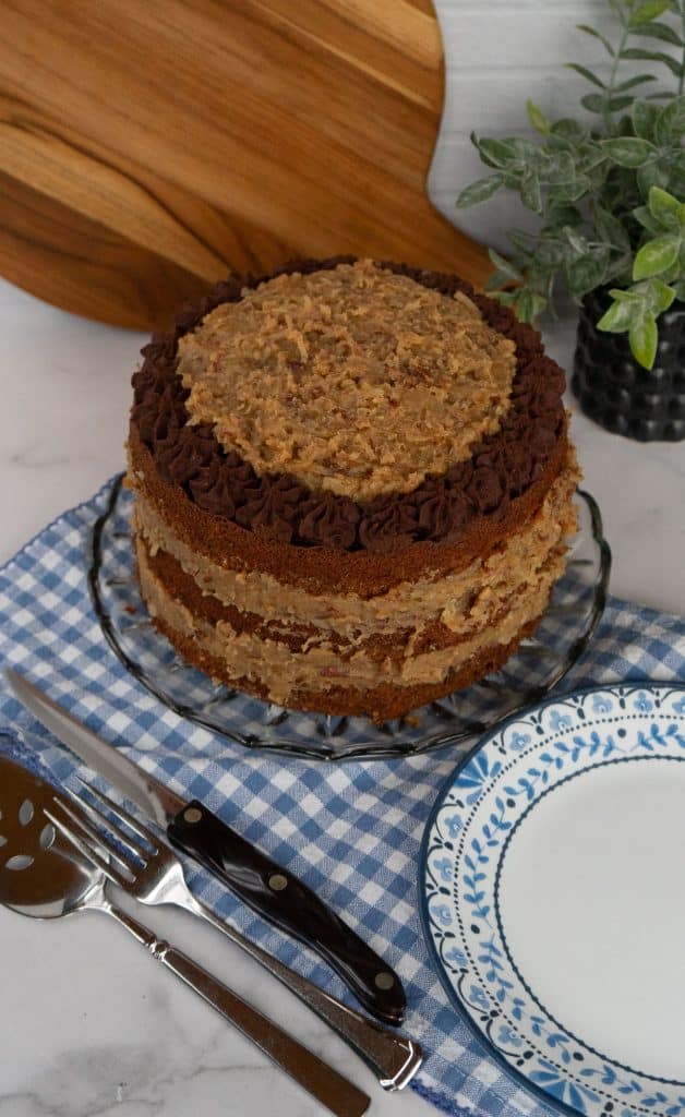 whole German Chocolate Cake with  on a glass cake plate with a blue and white gingham cloth underneath cake plate,  a blue trimmed plate is lower righthand corner
