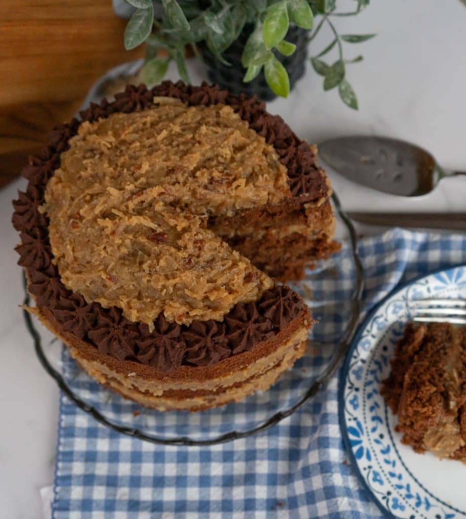 German Chocolate Cake with a slice cut out of it on a glass cake plate with a blue and white gingham cloth underneath cake plate, slice of cake is on a blue trimmed white plate cake server is in the upper right hand corner 