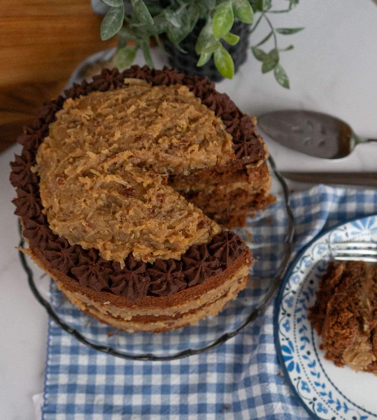German Chocolate Cake with a slice cut out of it on a glass cake plate with a blue and white gingham cloth underneath cake plate, slice of cake is on a blue trimmed white plate cake server is in the upper right hand corner
