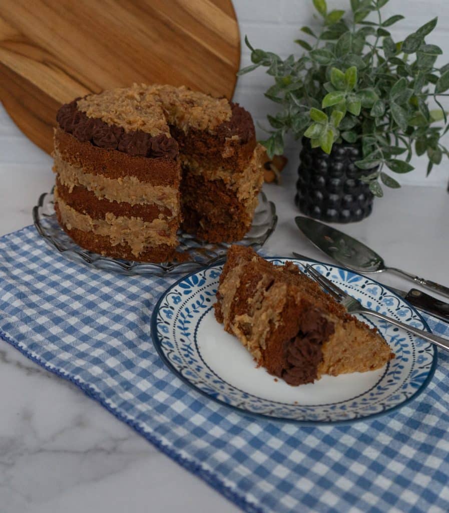 German Chocolate Cake with a slice cut out of it on a glass cake plate with a blue and white gingham cloth underneath cake plate, slice of cake is on a blue trimmed white plate cake server is in the upper right hand corner