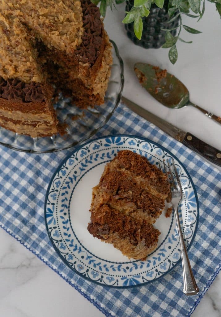 whole german chocolate cake on a glass plate with a piece taken out with a slice of cake on a plate with blue decorations on the edge with a fork on the plate and a knife and a cake server in the back ground a blue and white gingham cloth is underneath cake and cake plate