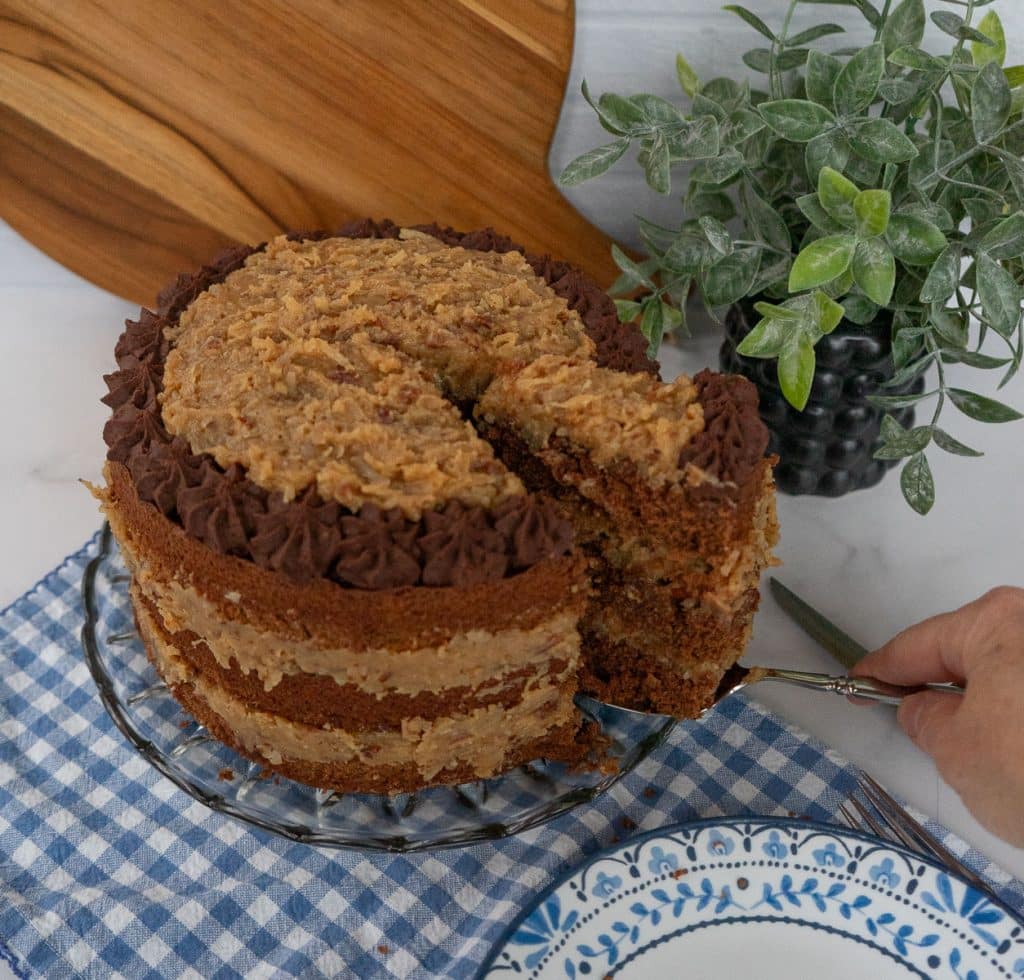 hand lifting a slice out of cut german chocolate cake with a blue rimmed plate in the lower right hand corner