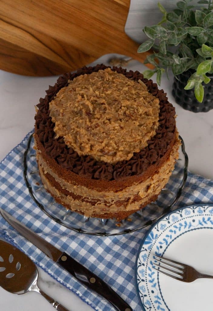 whole german chocolate cake on a glass plate sitting on a blue and white gingham cloth with a cake server and a knife in the lower left hand corner and a plate with a blue rim with a fork on it in the love right hand corner 
