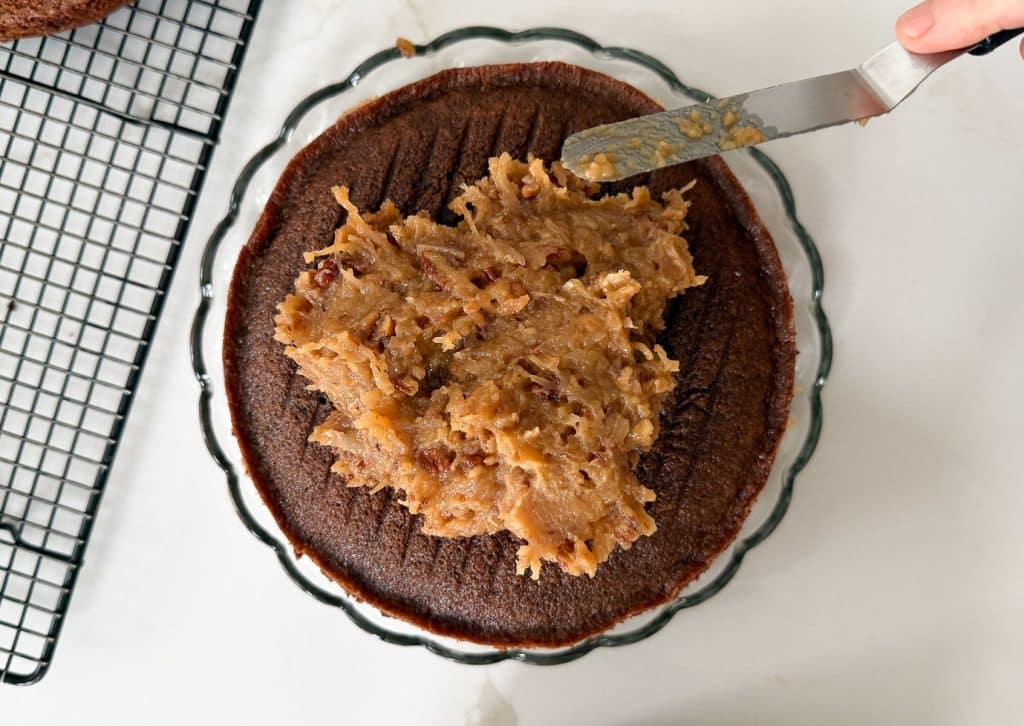 one layer of chocolate cake with pecan codonut frosting being spread with an off set spatula