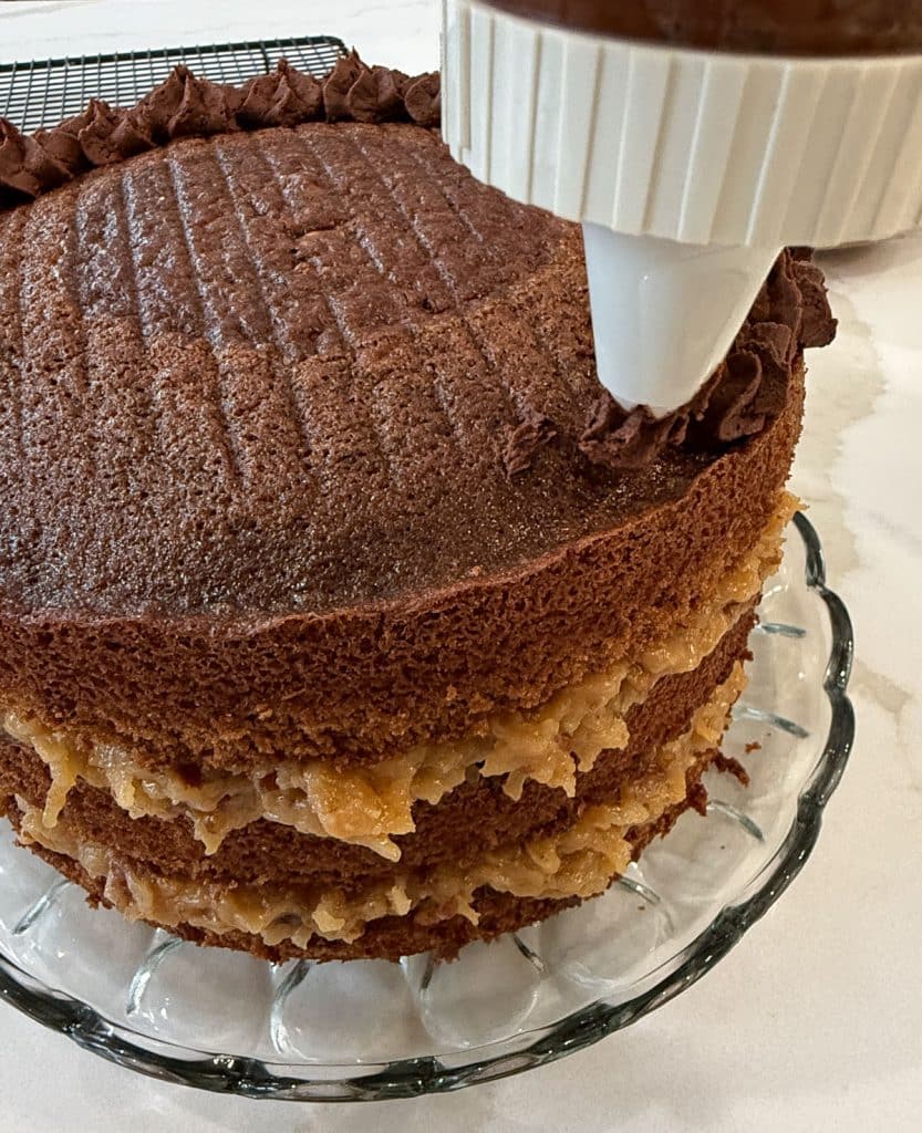 piping chocolate ganache around the top of the layered cake on a glass dish on a white counter top