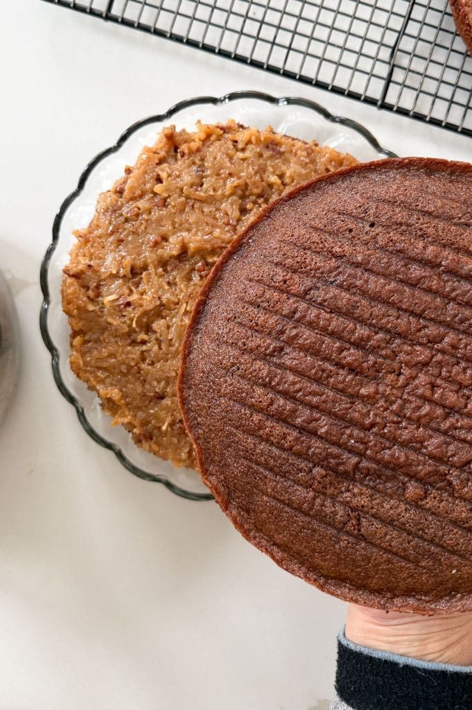 placing second layer on top of first layer of cake with coconut pecan filling in between cake is on a glass cakeplate on a white countertop