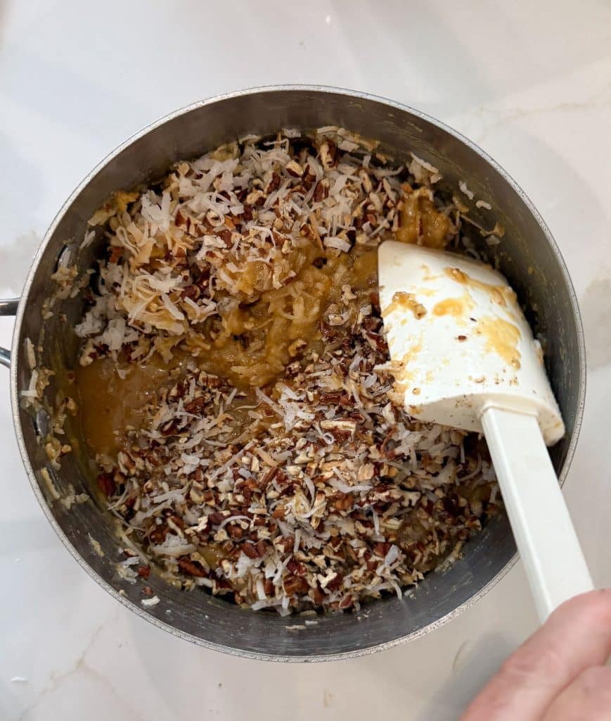folding coconut and pecans into cooked frosting on a white countertop with a white spatula