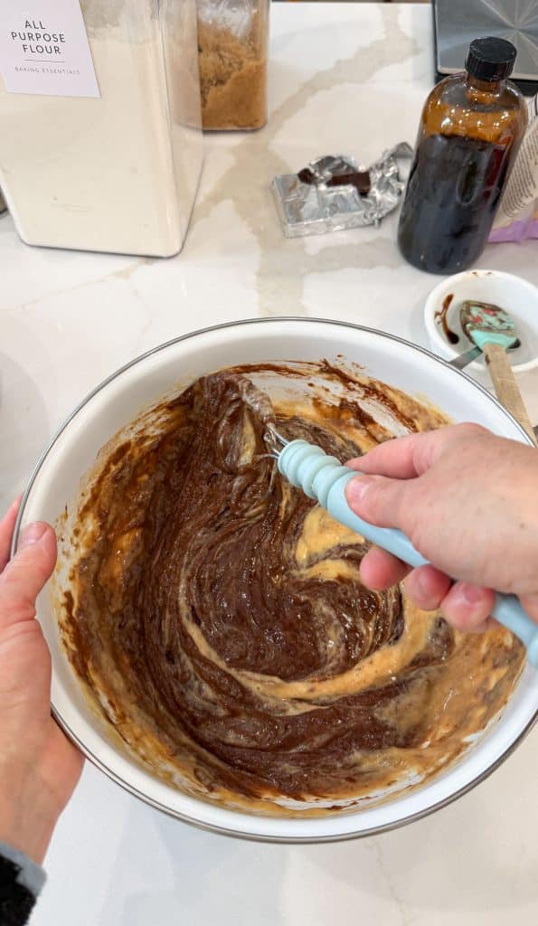 adding buttermilk egg mixture to chocolate mixture in a white bowl with a hand coming in from the right side whisking mixture together