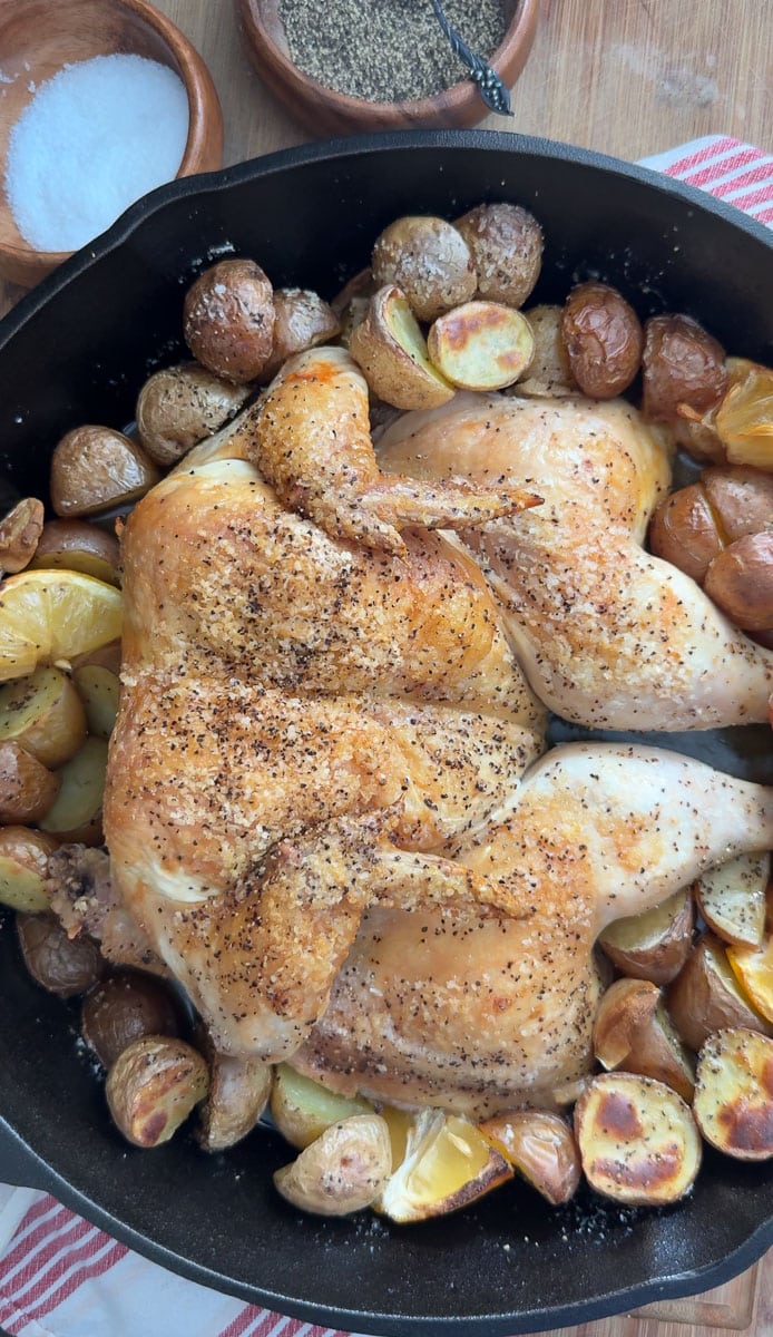 roast chicken in a cast iron skillet surrounded by roast potatoes, lemons and garlic