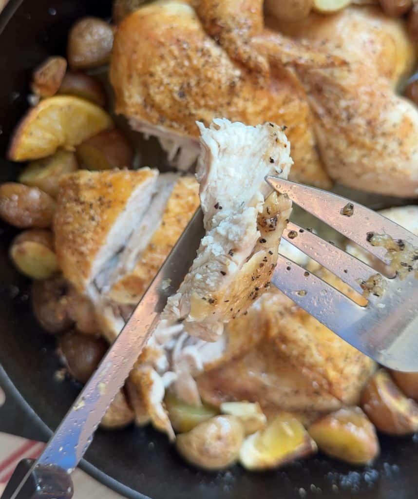roast chicken with a fork and knife cutting into the breast meat. chicken is in a cast iron skillet surrounded with potatoes, lemon, and garlic