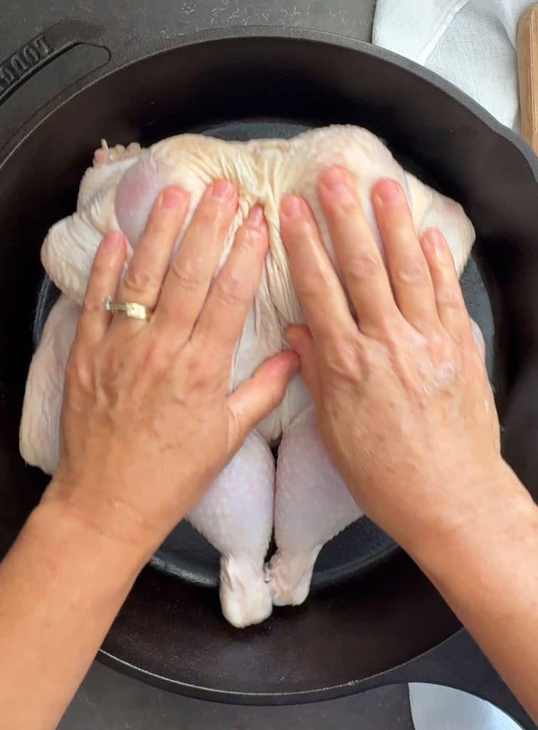 laying butterflied chicken in a cast iron skillet. hands are pressing chicken into the pan to flatten it out