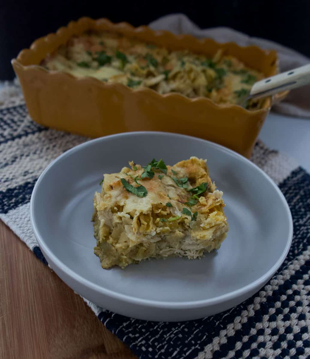 a piece of chicken enchiladas verde in a gray dish with the pan of enchiladas in the back ground