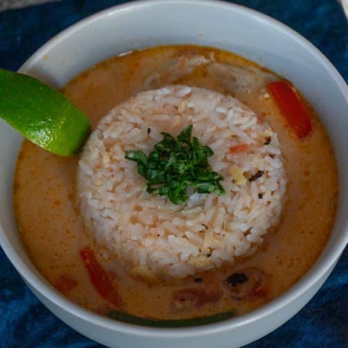 bowl of rice with thai coconut curry soup surrounding it with a lime wedge on the side of the bowl and it's garnished with cilantro