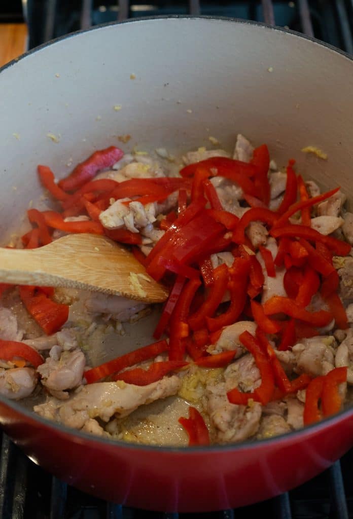 add sliced red pepper to pan and saute, a wooden spoon is moving ingredients in pan