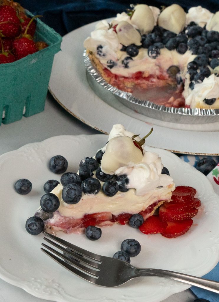 slice of rwb pie on a white plate with a fork on the plate the whole pie is in the back ground with a quart of fresh strawberries on a grey table