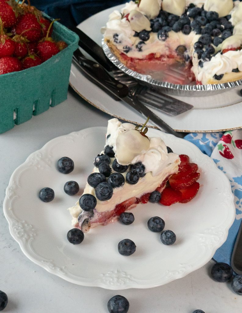 slice of rwb pie on a white plate with a fork on the plate the whole pie is in the back ground with a quart of fresh strawberries on a grey table